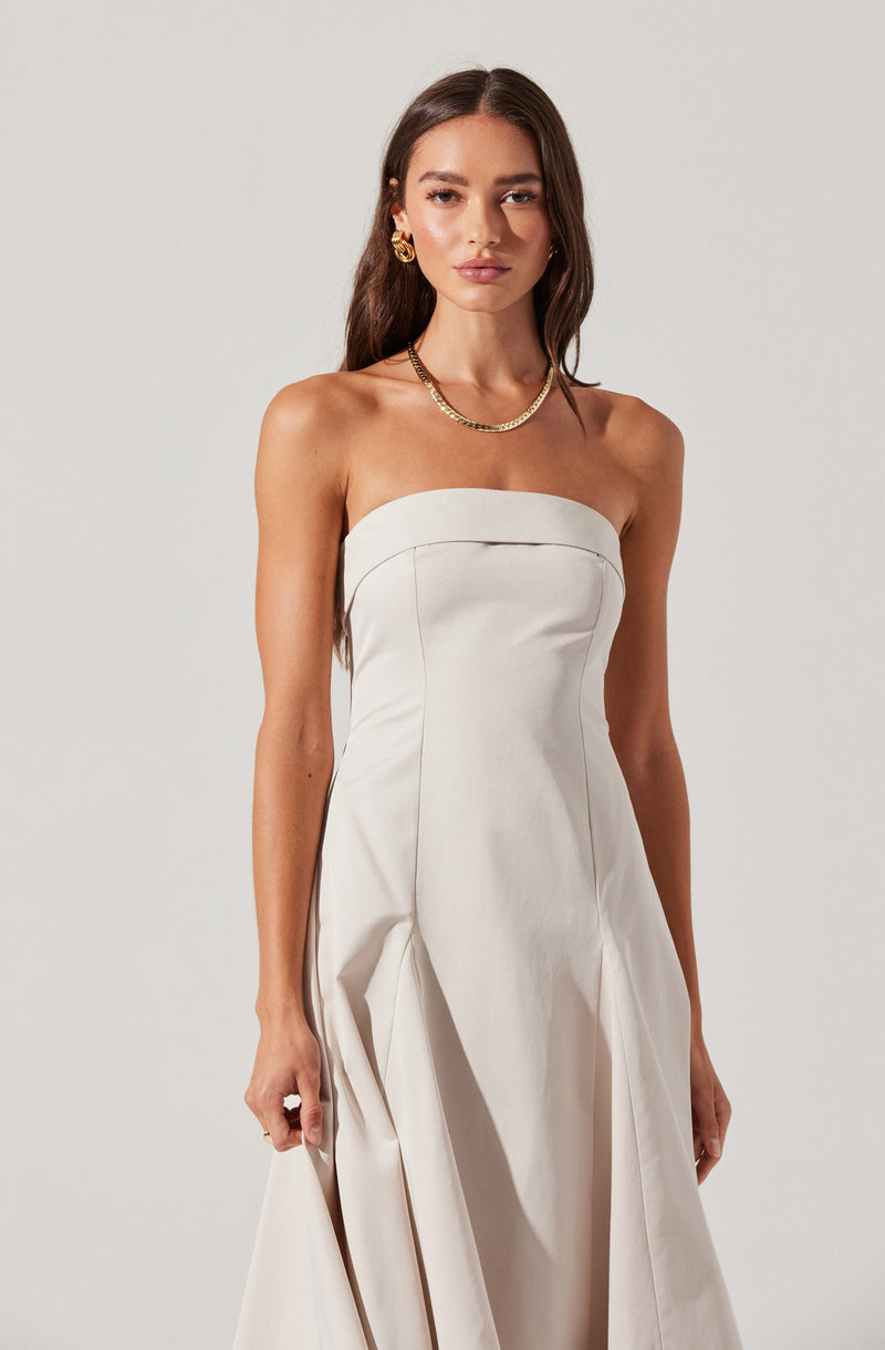 A woman is wearing a strapless white dress with a fitted bodice and pleated skirt. She accessorizes with gold jewelry, including a chain necklace and large earrings.