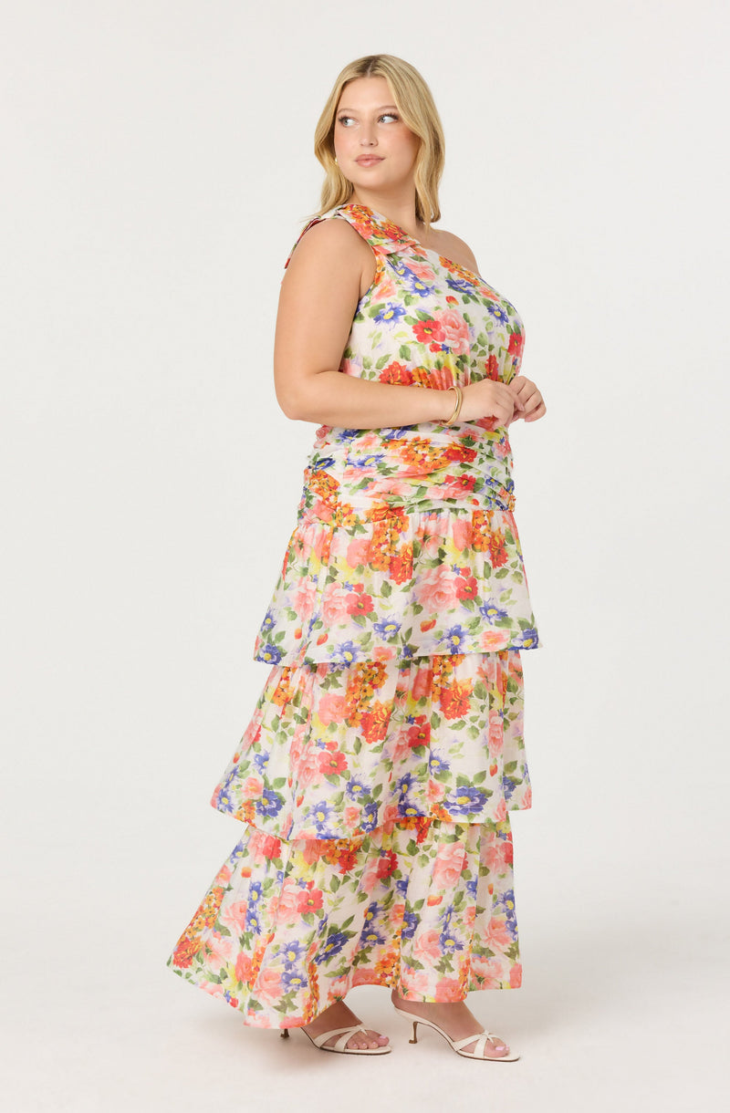 A woman is wearing a sleeveless, floral maxi dress with ruffled tiers. The dress features a colorful pattern with shades of pink, orange, purple, and green on a light background. She is also wearing open-toe, high-heeled sandals in a light color.