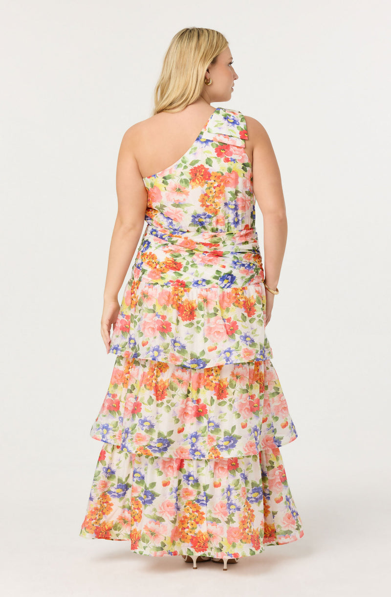 A woman is wearing a sleeveless, floral-patterned dress with multiple layers of ruffles. The dress has a one-shoulder design, with the shoulder on the left side. The fabric features a colorful flower print with shades of pink, orange, purple, green, and blue on a light background. She is accessorized with earrings and a bracelet.