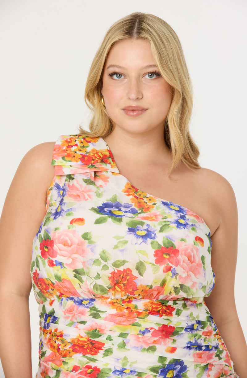 A woman is wearing a sleeveless, one-shoulder dress with a floral pattern featuring pink, red, purple, orange, and yellow flowers with green leaves on a white background. The dress has a ruffled detail on the shoulder.