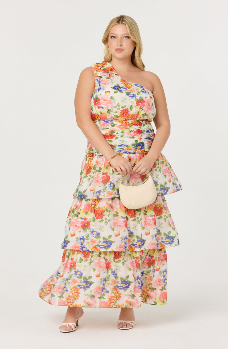 A woman is wearing a colorful floral dress with a one-shoulder neckline. The dress has a fitted bodice and a tiered, flowy skirt with multiple layers, all featuring a vibrant floral pattern. She is holding a small, light-colored handbag and wearing open-toe heels.