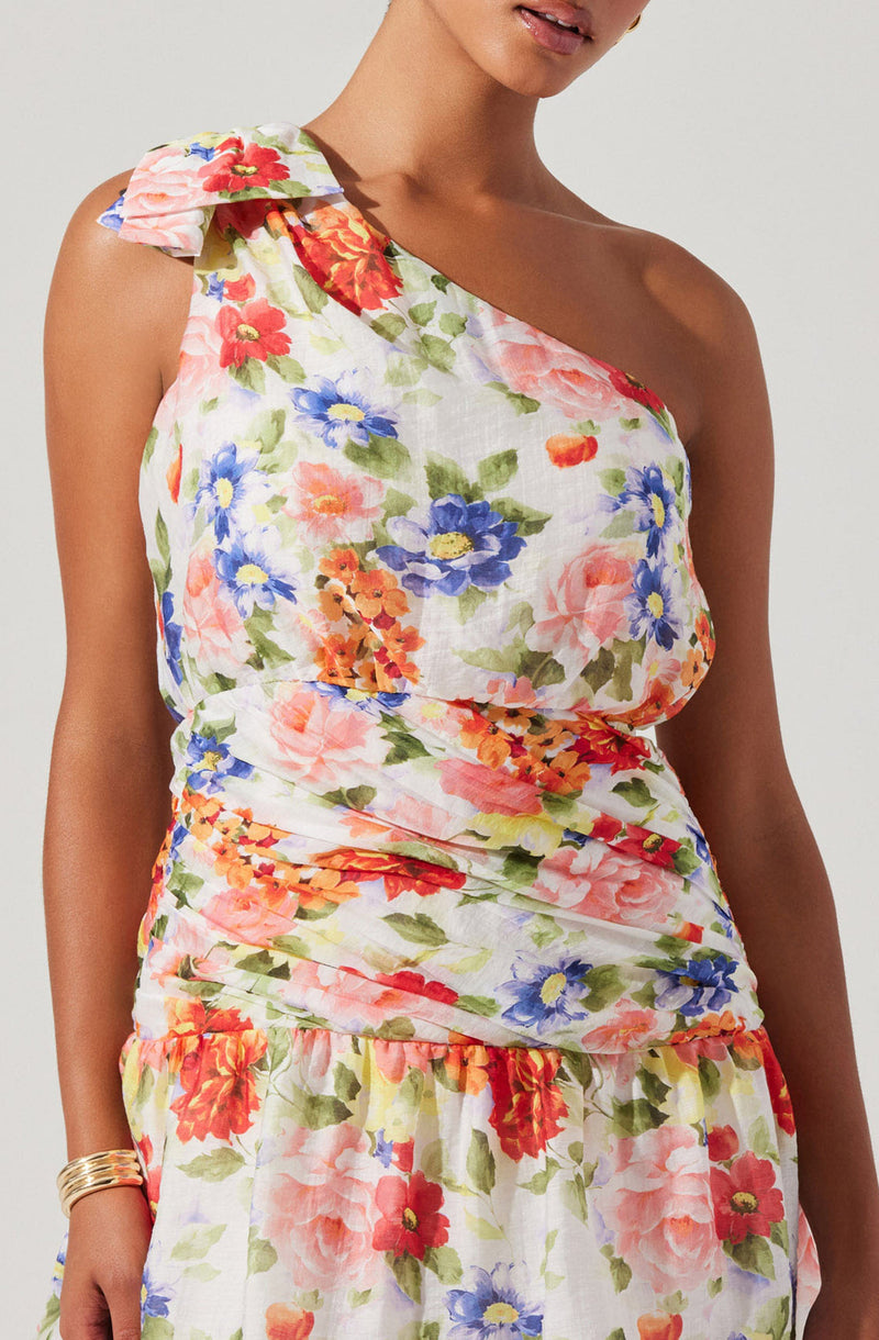 A woman wearing a sleeveless, floral print dress with a one-shoulder design featuring ruffle details on the shoulder strap. The dress has a fitted bodice with gathered fabric around the waist and a matching floral skirt with a loose, comfortable fit. The fabric has a white background with colorful flowers in shades of pink, red, blue, orange, and green. She is also wearing a gold bracelet on her right wrist.