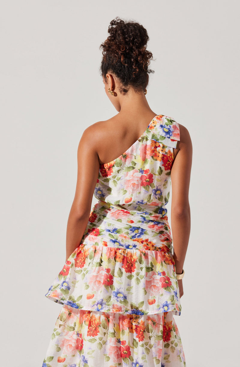A woman is wearing a sleeveless, one-shoulder dress with a vibrant floral pattern in shades of pink, red, purple, blue, and green. The dress features a ruffled detail at the shoulder and has a tiered skirt with a ruffled hem.