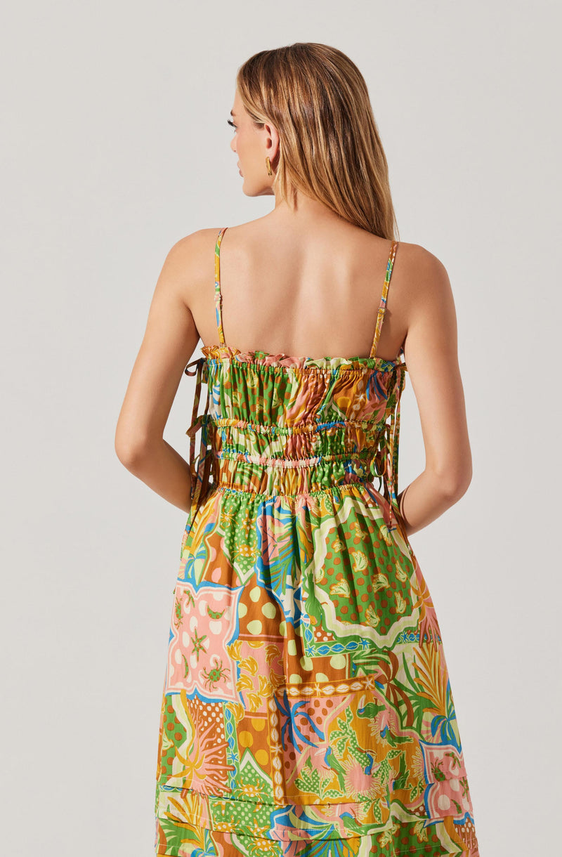 A woman is wearing a colorful, patterned sundress with thin spaghetti straps. The dress features a mix of bright colors, including green, pink, orange, blue, and yellow, with various abstract and floral designs. The top portion is gathered with elastic, creating a ruched effect, and the skirt is flowing with a vibrant, patchwork-like pattern.