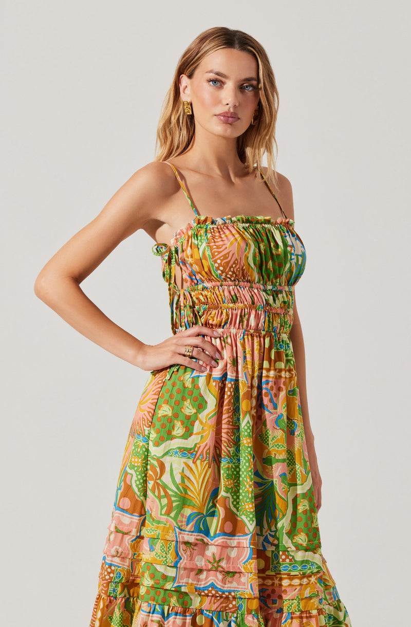 A woman is wearing a colorful, patterned sundress with thin, adjustable spaghetti straps. The dress features a smocked bodice with ruffled edges and a flowing skirt decorated with various vibrant, tropical-inspired prints including leaves, flowers, and abstract designs in shades of orange, green, pink, blue, and yellow.