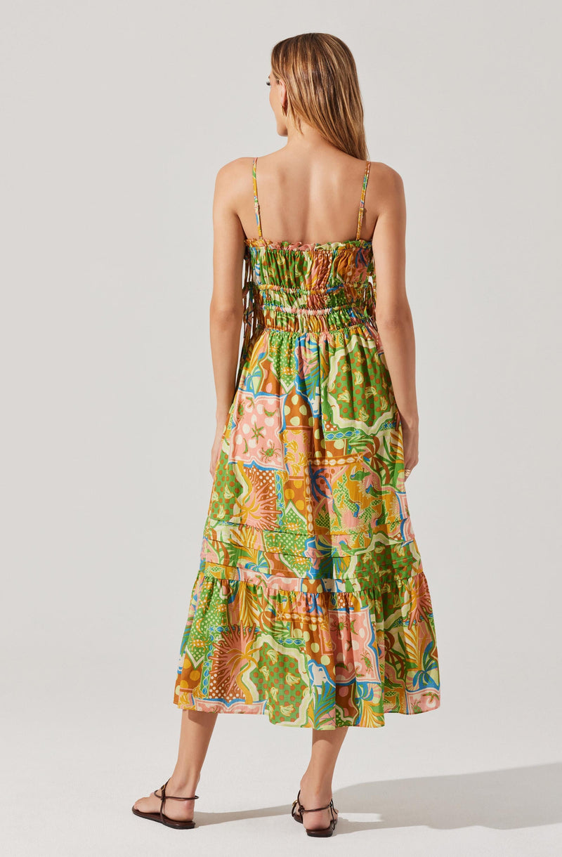 A woman is wearing a colorful, sleeveless, ankle-length dress with thin straps. The dress features a vibrant, abstract pattern with shades of green, pink, orange, and blue. The fabric appears lightweight and flowy, suitable for warm weather.