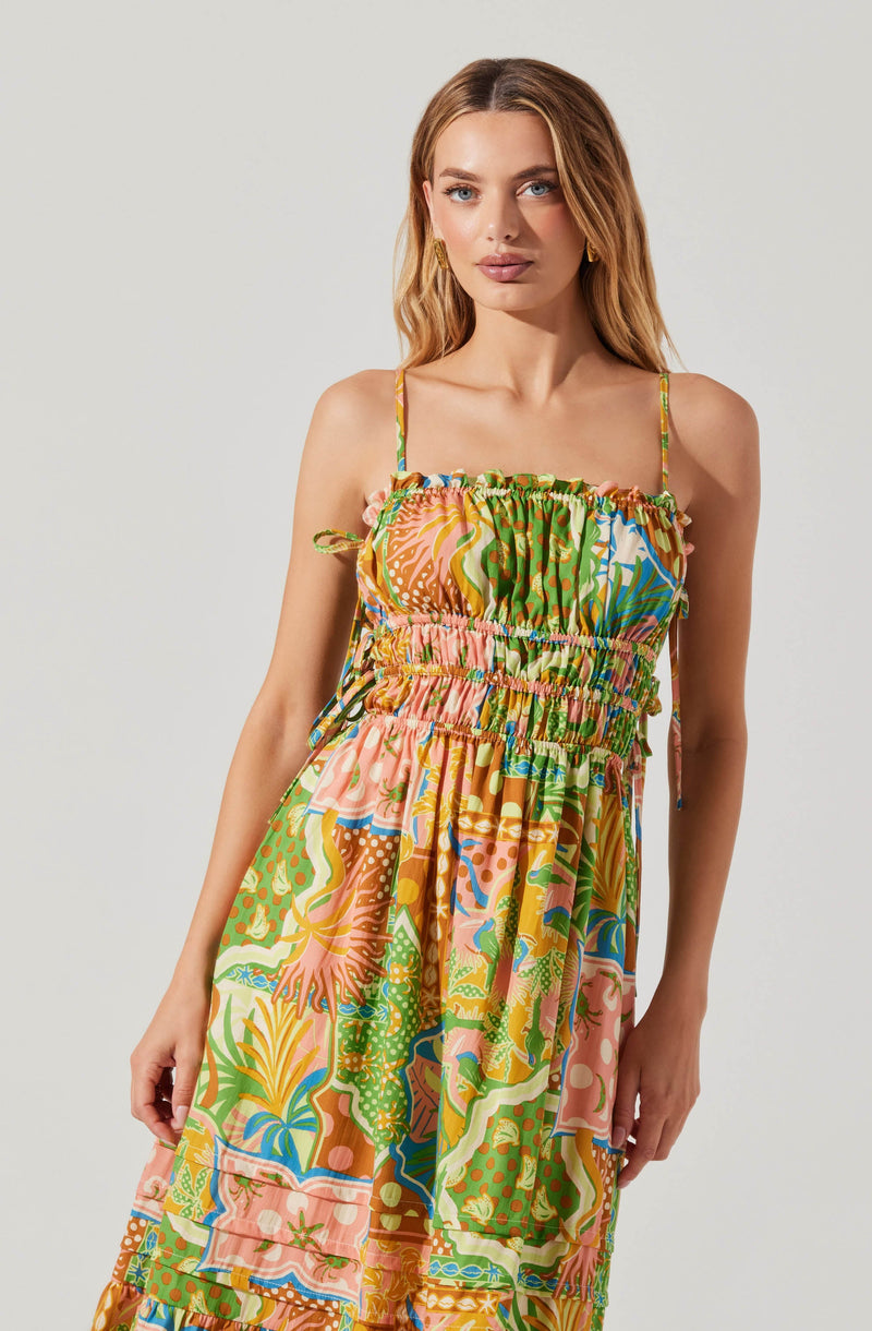 A woman wearing a colorful, patterned dress with thin spaghetti straps. The dress features an elastic, smocked bodice and a flowing skirt with vibrant, abstract and floral designs in shades of green, pink, yellow, blue, and orange.