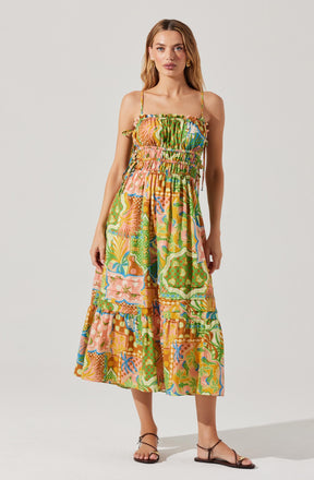 A woman is wearing a colorful, sleeveless, ankle-length dress with adjustable spaghetti straps. The dress features a vibrant, multicolored pattern with various abstract and floral designs, and has a smocked waistline for a fitted look. The fabric appears lightweight and flowy. She is also wearing simple, flat, dark-colored sandals.