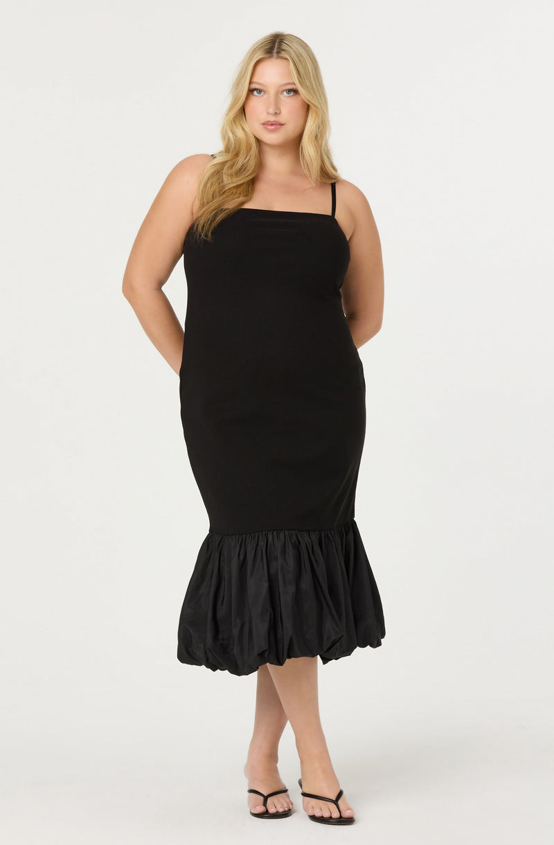 A woman is wearing a black dress with thin spaghetti straps, fitted through the bodice and hips, with a gathered, puffy hem at the bottom. The dress has a sleek, smooth texture with a ruffled, puffed section at the lower hem. She is also wearing black high-heeled sandals.