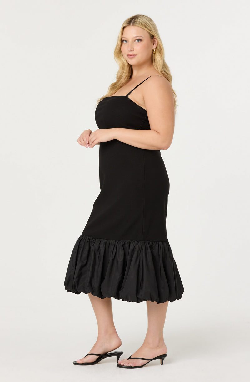 A woman is wearing a black sleeveless dress with thin spaghetti straps. The dress is fitted at the waist and has a ruffled, gathered hem that extends to mid-calf. She is also wearing black open-toe high-heeled sandals.