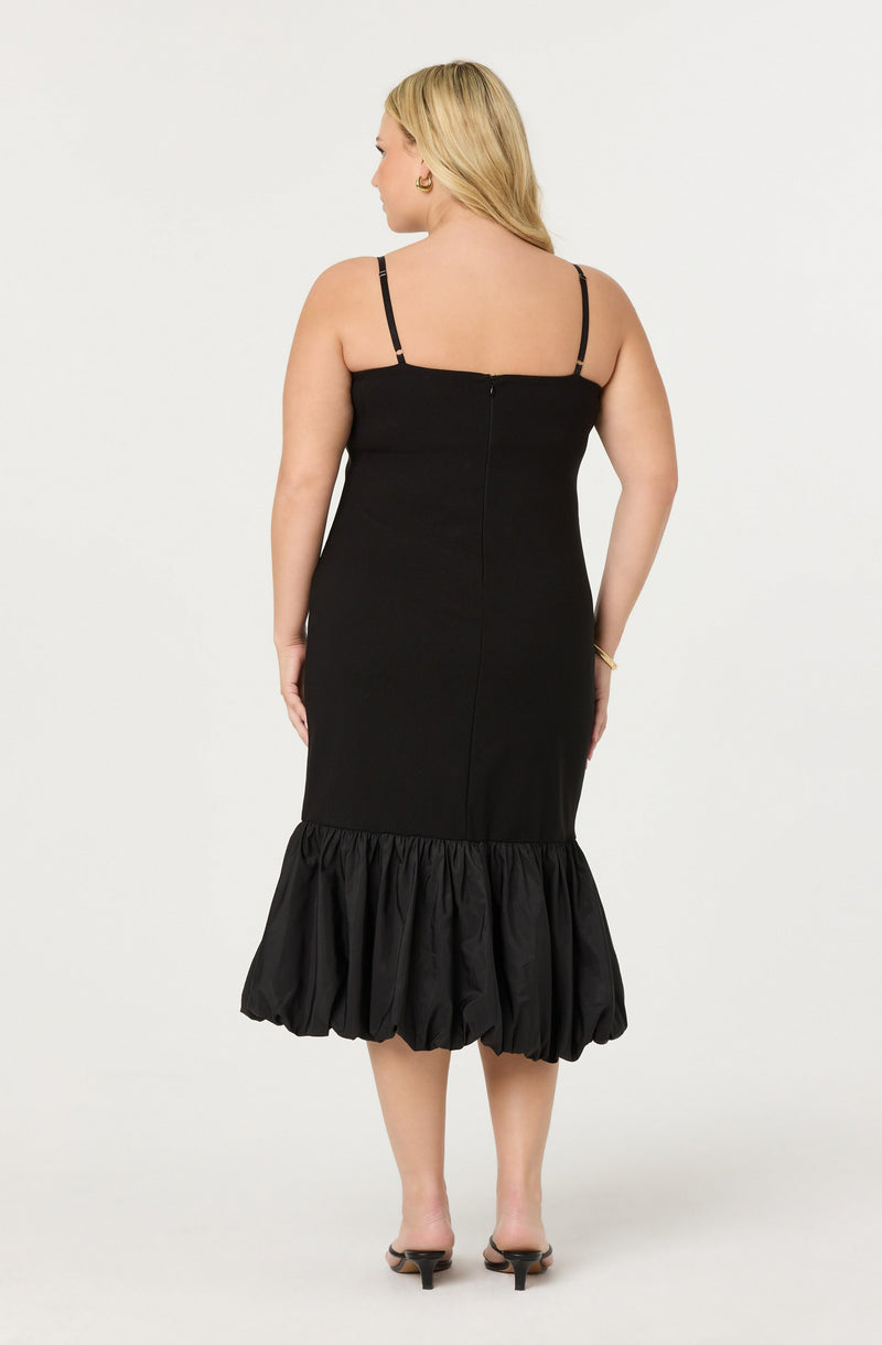 A woman is wearing a sleeveless black dress with thin spaghetti straps. The dress has a fitted bodice and features a ruffled, gathered hem at the bottom that extends below the knees. She is also wearing black open-toe heeled sandals.
