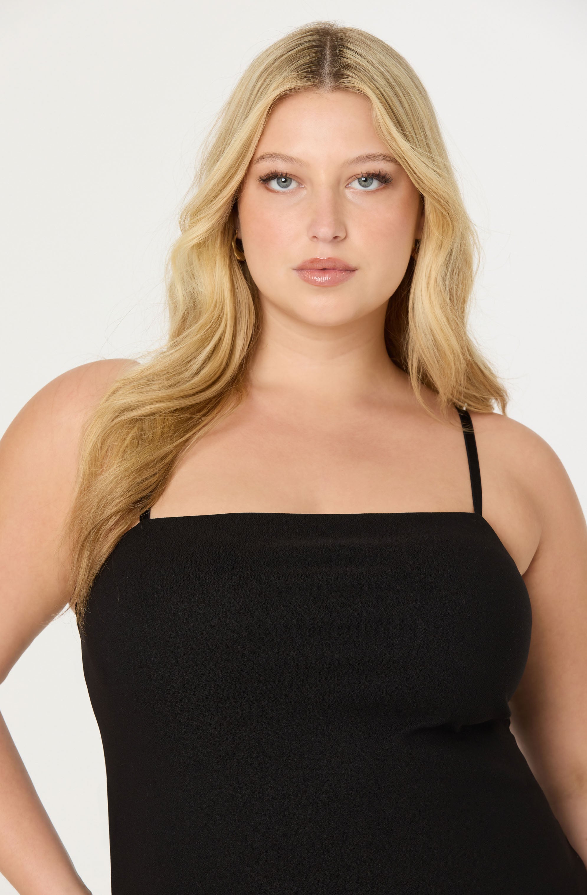A woman with long, wavy blonde hair wears a black spaghetti strap top. The top fits snugly and has a straight neckline. She is also wearing small hoop earrings.