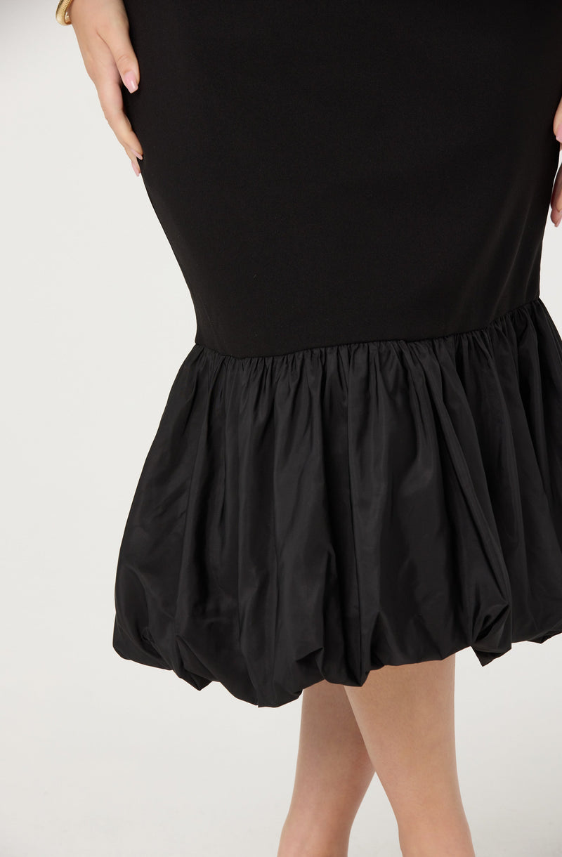 A close-up of a person wearing a black dress with a fitted upper part and a gathered, voluminous lower part made from shiny, ruffled fabric. The person's left hand is resting on their hip.