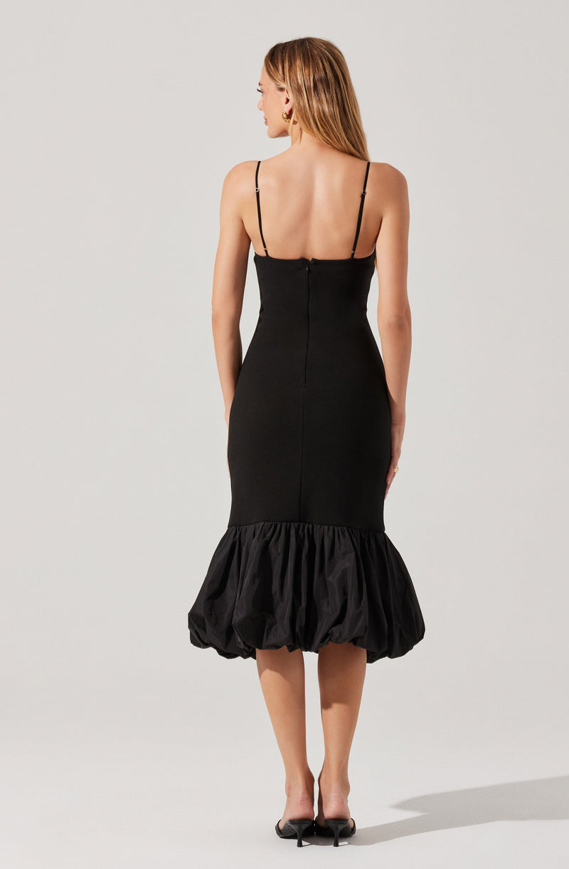 A woman is wearing a fitted black dress with thin spaghetti straps. The dress is sleek through the upper and midsection, then features a voluminous, gathered bubble hem at the bottom, adding a dramatic and stylish touch. The back of the dress has a concealed zipper closure. The woman is also wearing black high-heeled shoes.
