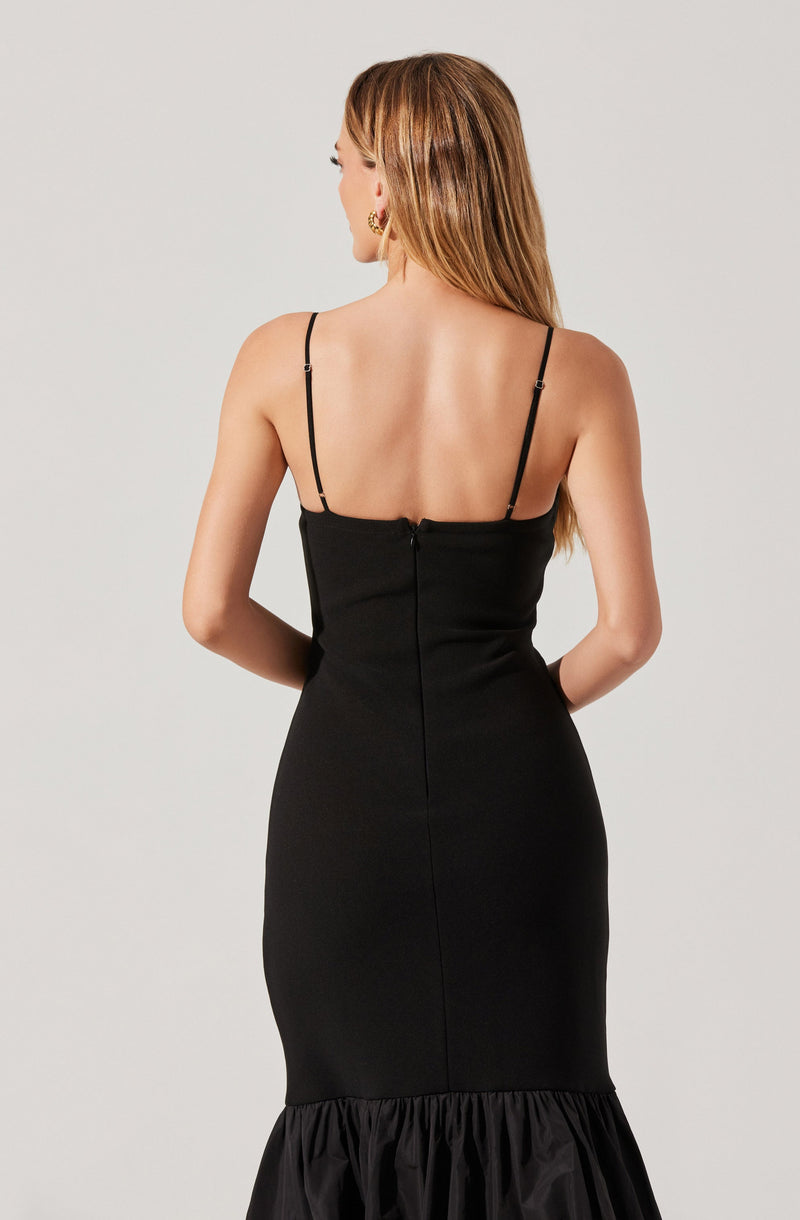 A woman is wearing a fitted black dress with thin spaghetti straps. The dress has a sleek design with a zipper at the back and a ruffled hem at the bottom.