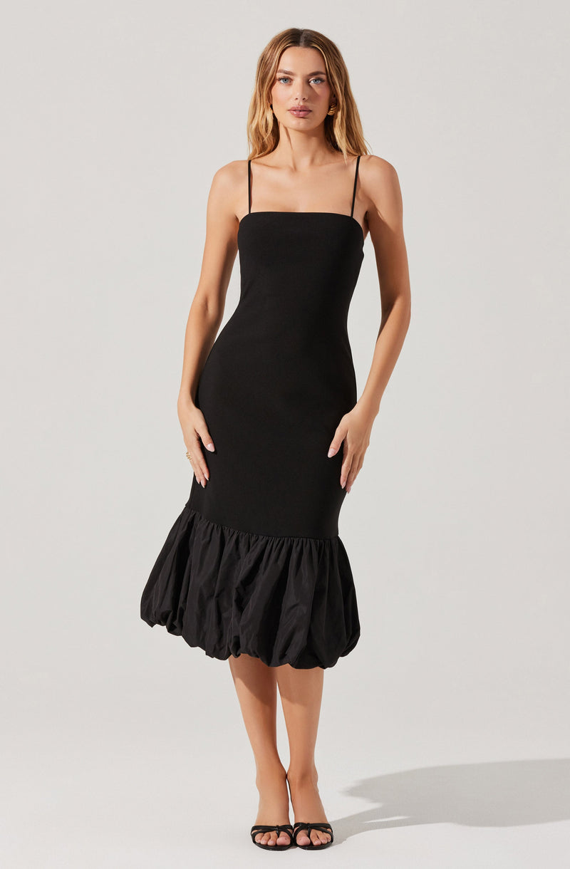 A woman is wearing a black sleeveless dress with thin spaghetti straps. The dress has a fitted bodice and a gathered, voluminous bubble hem at the bottom. She is also wearing black open-toe high-heeled shoes.