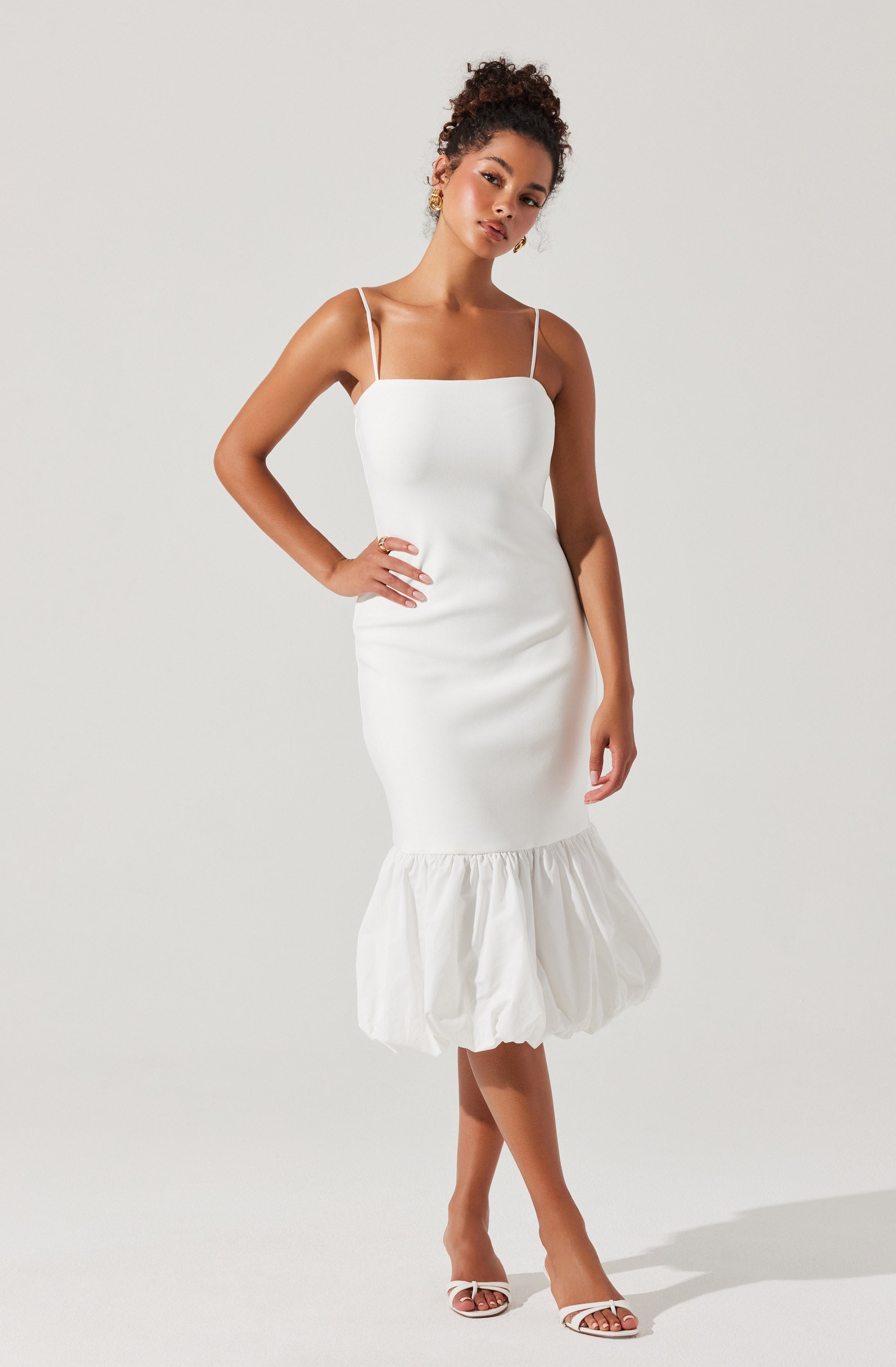 A woman is wearing a white, fitted dress with thin spaghetti straps. The dress has a straight neckline and a ruched section around the hem, which features a voluminous, gathered bubble skirt detail. She is also wearing white open-toe high heels.