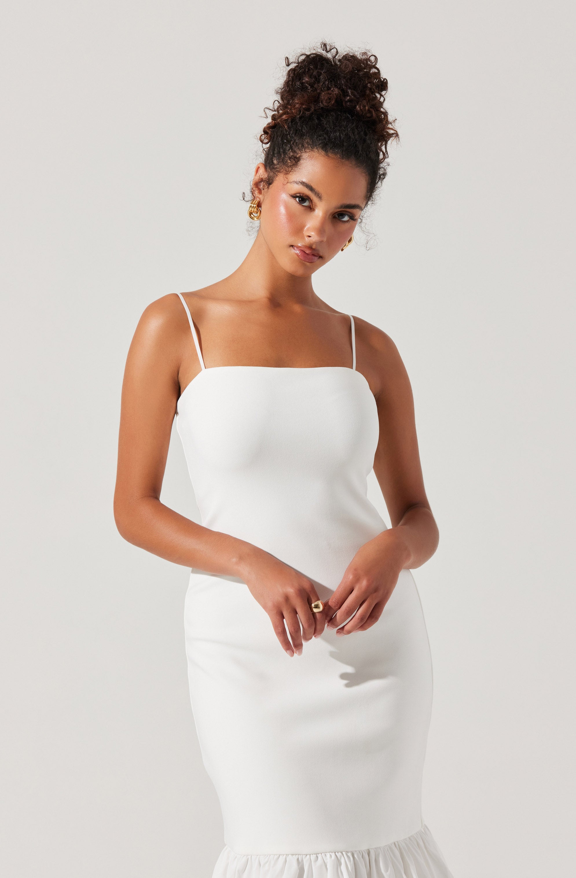 A woman is wearing a fitted, sleeveless white dress with thin spaghetti straps. The dress has a smooth, sleek fabric and a ruffled hem at the bottom.
