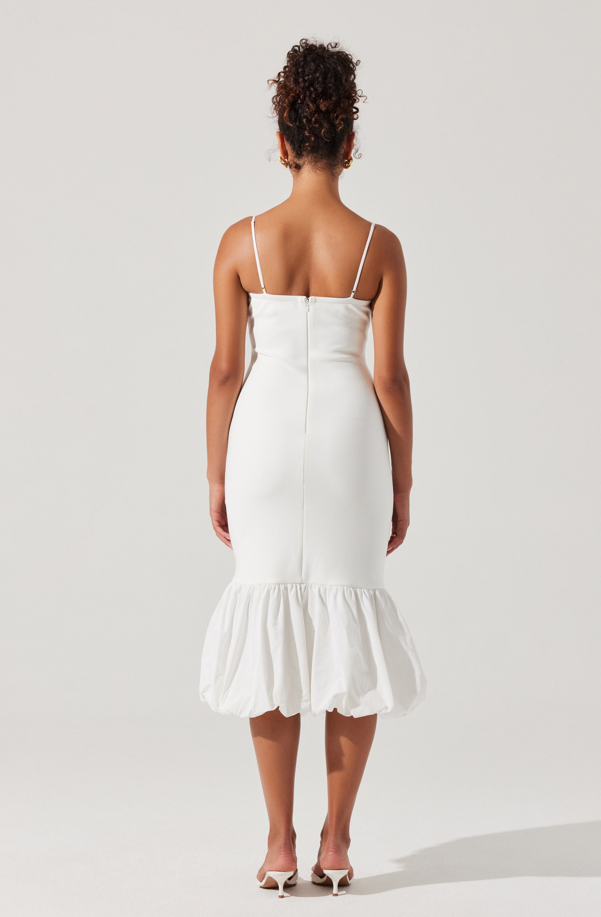 A woman is wearing a white dress with thin spaghetti straps, a fitted bodice, and a midi-length skirt that features a gathered, ruffled hem at the bottom. She is also wearing white high-heeled shoes.