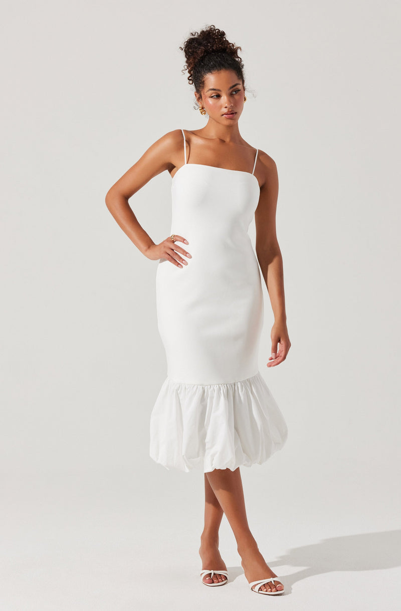A woman is wearing a white dress with thin spaghetti straps. The dress is fitted through the torso and hips, then features a ruffled, gathered hem that falls below the knees. She is also wearing white heeled sandals.