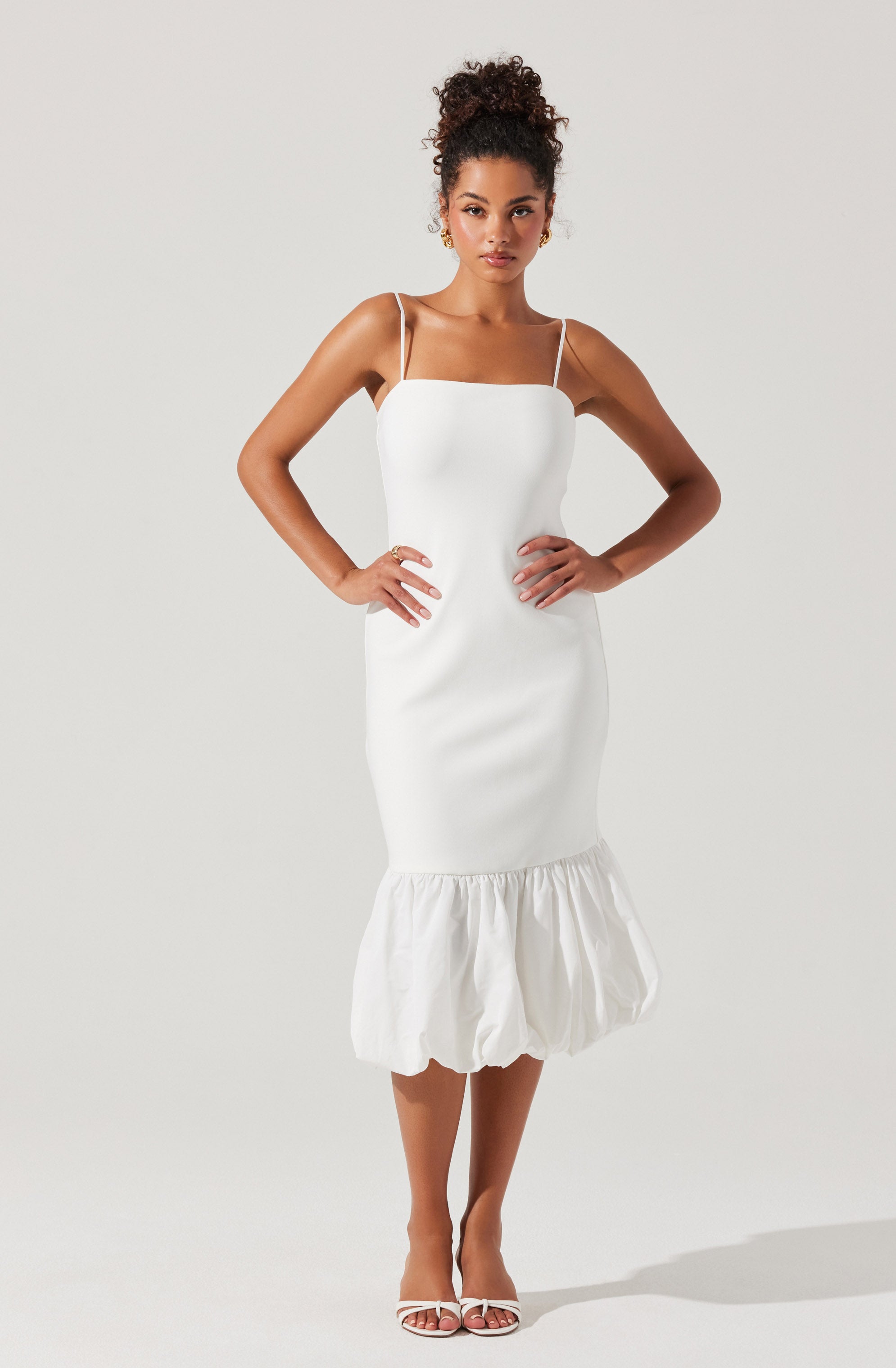 A woman is wearing a white sleeveless dress with thin spaghetti straps. The dress has a fitted bodice that extends to a slightly flared, gathered ruffle at the hem, which is mid-calf length. She is also wearing white open-toe high-heeled sandals with thin straps.