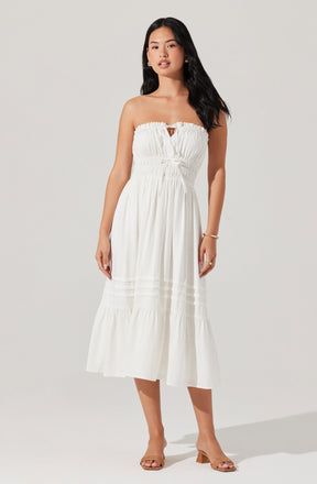 A woman is wearing a strapless white maxi dress with a ruched bodice and a small keyhole opening at the center of the neckline. The dress has a fitted waist and a flowy skirt with horizontal seam details. She is also wearing open-toed, block-heeled sandals in a neutral tone.
