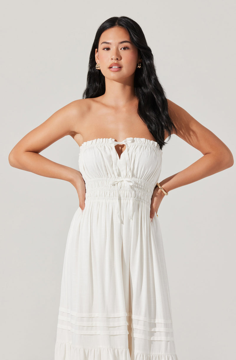 A woman is wearing a strapless, white, lightweight jumpsuit with a gathered, elasticized top featuring a small keyhole cutout in the center. The jumpsuit has a flowing, wide-leg design with horizontal pleats near the hem.