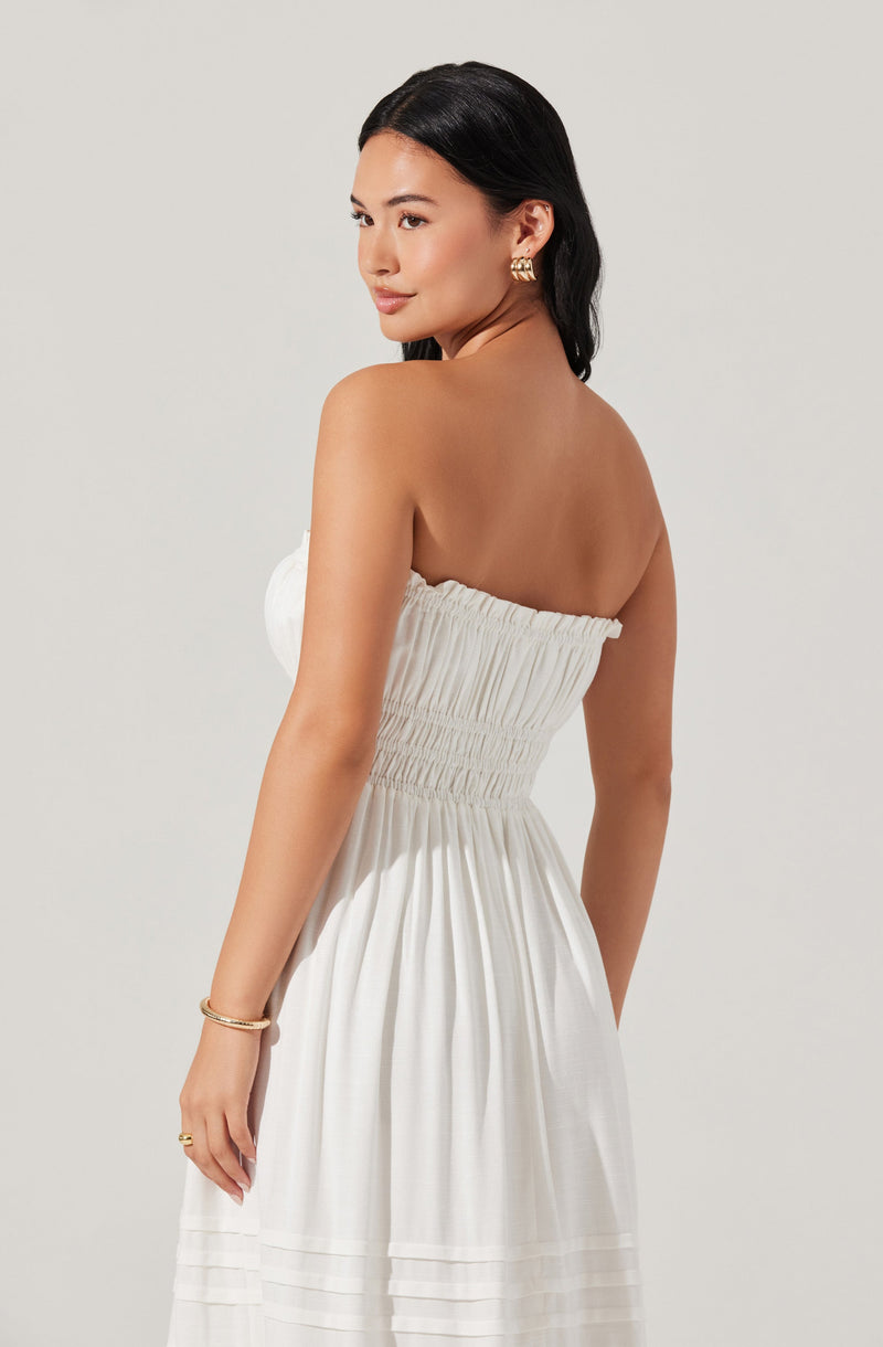 A woman is wearing a strapless, white dress with a ruched bodice that gathers at the waist. The dress has a flowing skirt with horizontal pleats near the hem. She accessorizes with gold jewelry, including earrings, a bracelet, and rings.
