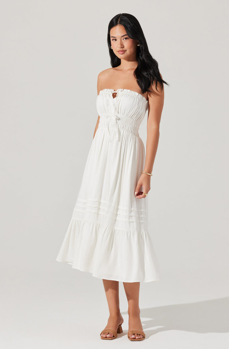 A woman is wearing a strapless white maxi dress with a gathered bust and a small keyhole cutout at the center. The dress has a fitted waist with drawstring detailing and a tiered, flowing skirt that extends to her mid-calf. She is also wearing open-toed beige high-heeled sandals and a gold bracelet on her left wrist.