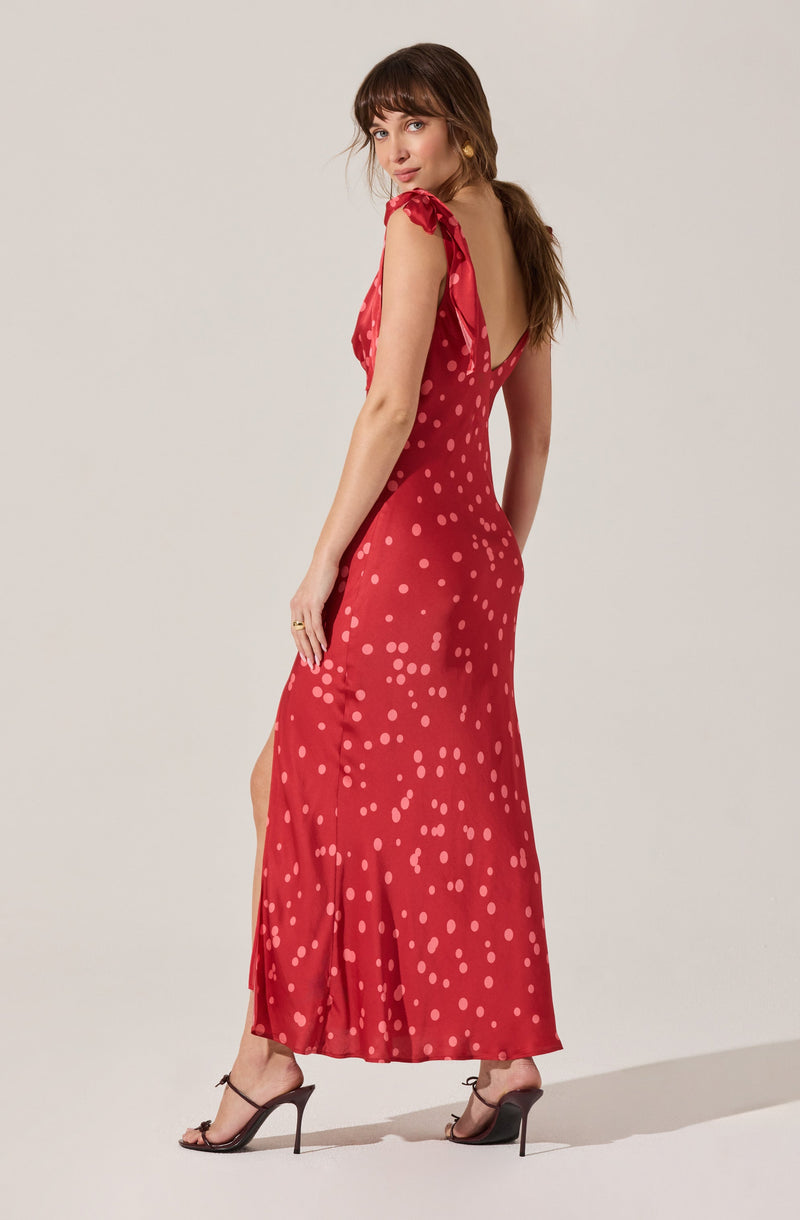 A woman is wearing a red maxi dress with a polka dot pattern. The dress features ruffled shoulder straps, a low V-back, and a slit on the side. She is also wearing black high-heeled sandals and has her hair styled in loose waves.