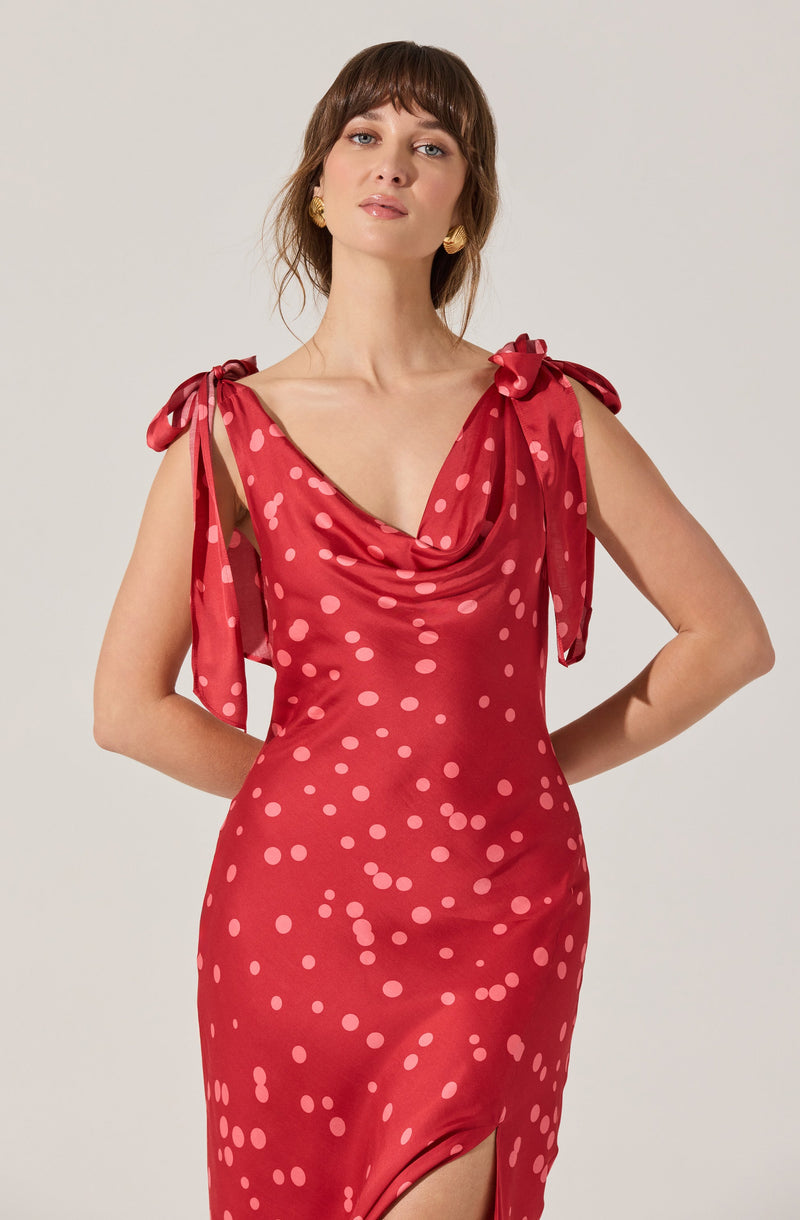 A woman is wearing a sleeveless, red dress with pink polka dots. The dress features a draped cowl neckline and shoulder ties that form bows. The dress has a fitted silhouette with a slight slit at the hem.