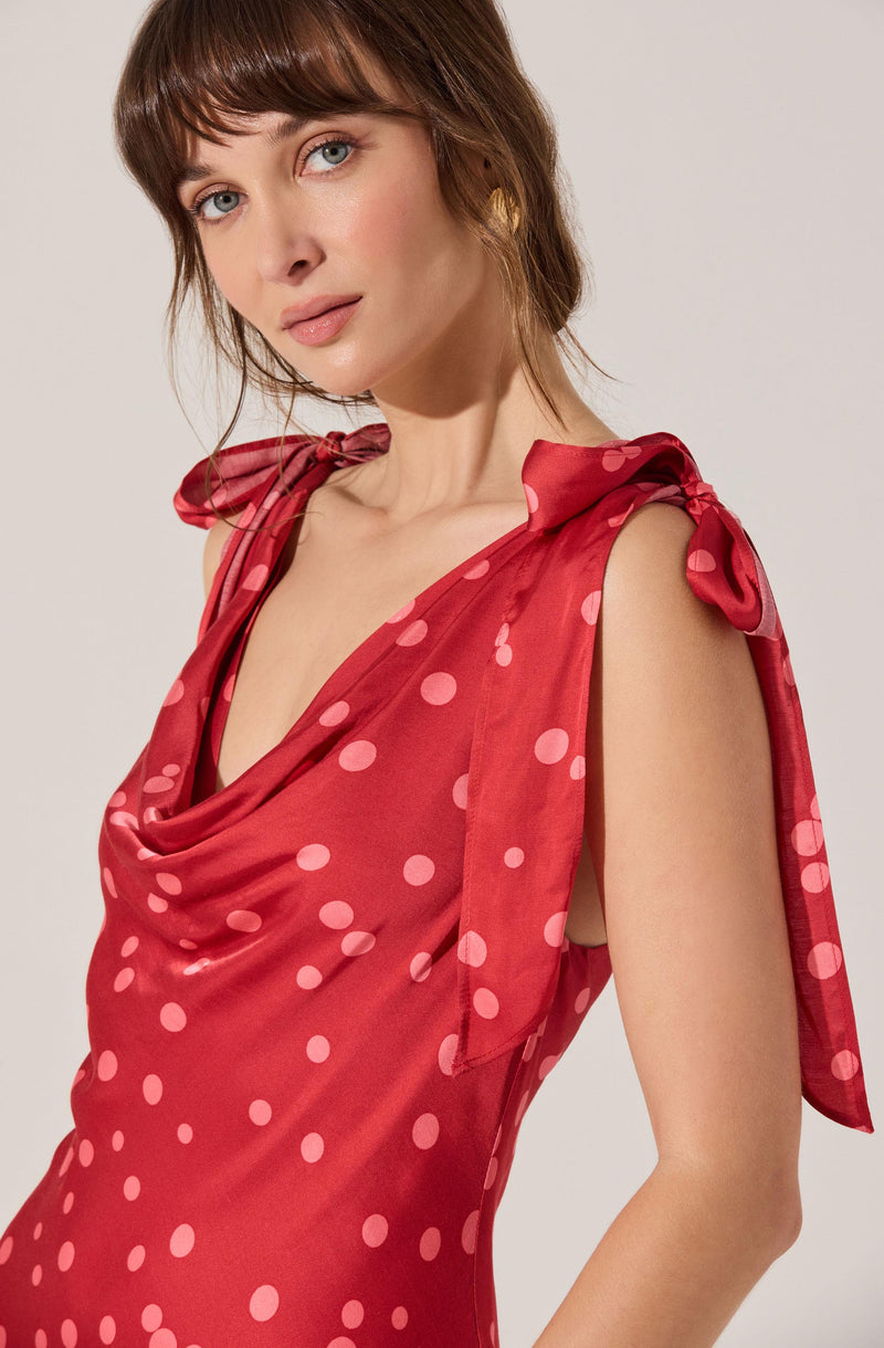 A woman wearing a sleeveless red satin dress with pink polka dots. The dress features a draped neckline and shoulder straps tied into bows.