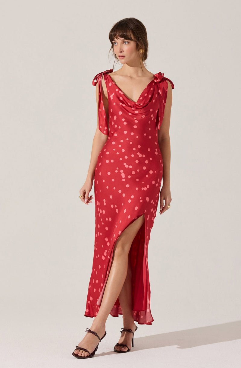 The dress is a sleeveless, full-length gown in a vibrant red color with a pattern of pinkish-white polka dots. It features a draped cowl neckline and shoulder ties that form small bows. The dress has a high slit on the left side, revealing the leg, and the fabric appears silky with a slight sheen. The overall style is elegant and suitable for formal occasions.
