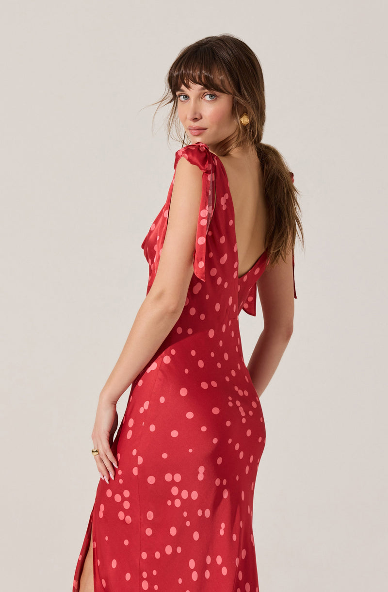 A woman is wearing a red, floor-length dress with a slit on the side. The dress features a pattern of pink polka dots, with the dots becoming more scattered towards the bottom. The dress has a deep V back and ruffled, short sleeves with tie details on the shoulders.