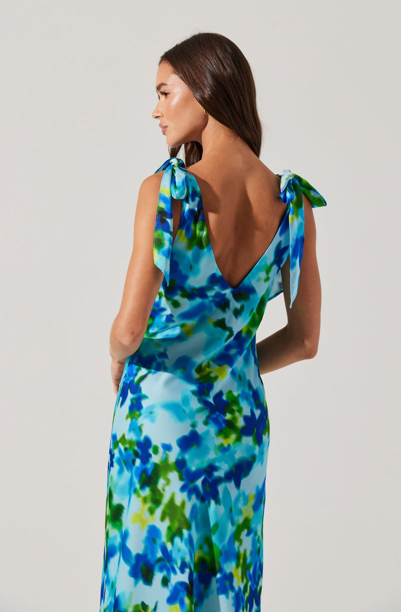 A woman is wearing a sleeveless, floor-length dress with a vibrant, abstract pattern in shades of blue, green, and white. The dress features shoulder ties that form small bows, and it has a deep V-back design.