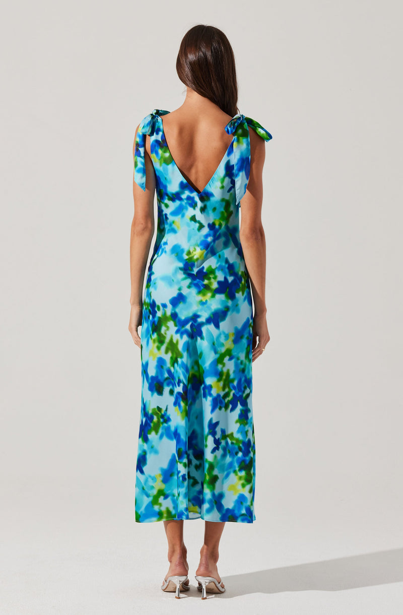 A woman is wearing a sleeveless, ankle-length dress with a V-shaped back. The dress features shoulder straps tied into bows and has a vibrant, multicolored abstract floral pattern in shades of blue, green, and white. She is also wearing white heeled sandals.