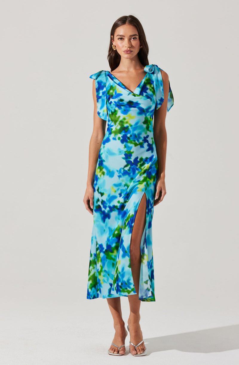 A woman is wearing a long, flowing dress with a vibrant, abstract floral pattern in shades of blue, green, and light blue. The dress features bow-tied straps on the shoulders and a high slit on one side, revealing her leg. She is also wearing open-toed, strappy heels.