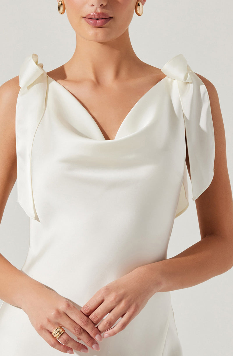 The dress is made of a smooth, ivory satin fabric with a subtle sheen. It features a sleeveless design with shoulder details that are tied into bows, creating a soft, feminine look. The neckline is draped in a cowl style, adding elegance to the overall design.