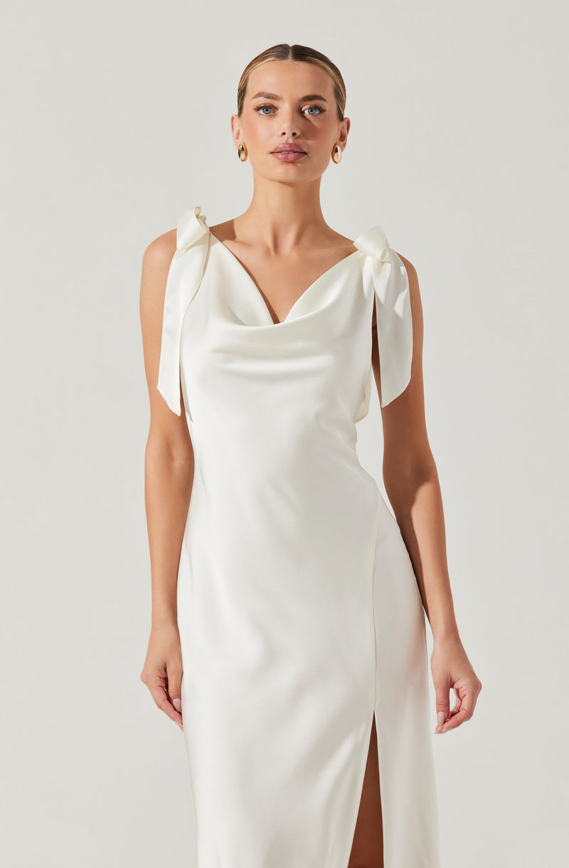A woman is wearing a sleeveless, white satin dress with a cowl neckline. The dress features shoulder straps tied into large bows, adding a decorative touch. The skirt has a slit on one side.