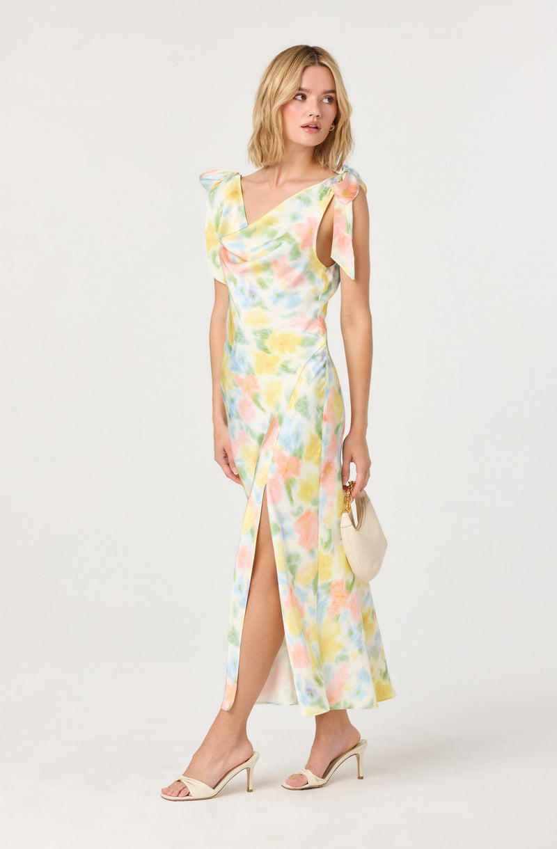 A woman is wearing a sleeveless, floor-length dress with a colorful pastel watercolor pattern. The dress has a V-neckline, ruffled shoulder details, and a front slit that reveals her leg. She is holding a small beige clutch bag and wearing nude high-heeled sandals.