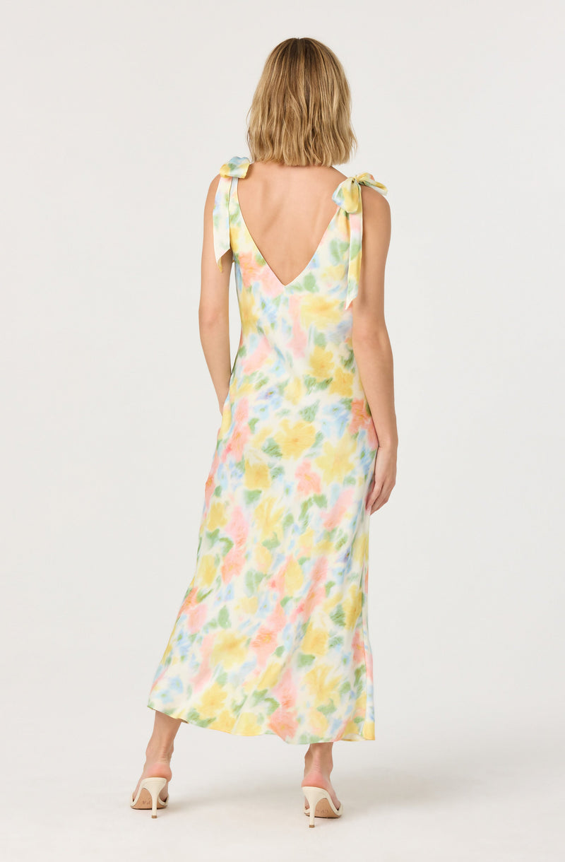 A woman is wearing a sleeveless, ankle-length dress with a watercolor floral pattern in pastel shades of yellow, pink, green, and blue. The dress features shoulder straps tied into bows on each shoulder and a deep V-shaped back. She is also wearing light-colored high-heeled shoes.