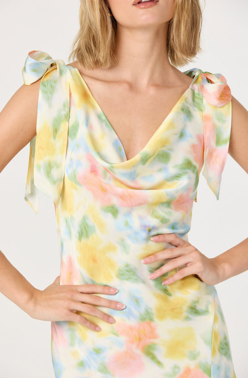 A woman is wearing a sleeveless dress made from a lightweight, satin-like fabric with a colorful tie-dye pattern featuring shades of yellow, pink, green, and blue. The dress has a draped neckline and shoulder ties that form bows on each shoulder.