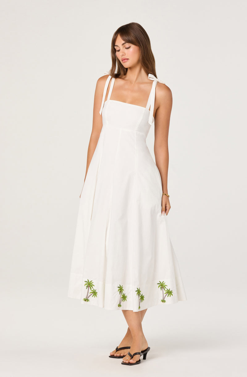 Woman wearing a white sleeveless dress with shoulder straps tied into bows and embroidered palm trees at the hem.