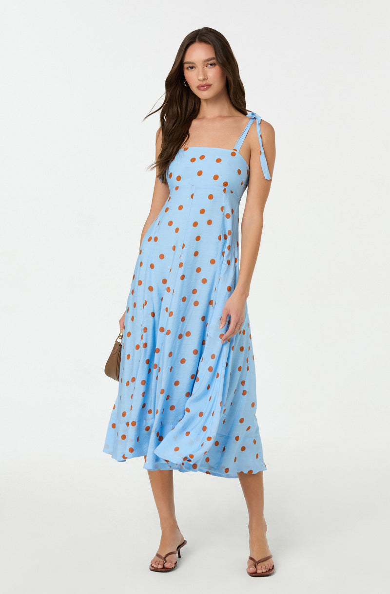 A woman is wearing a light blue, sleeveless, midi-length dress with thin shoulder straps tied into bows on her shoulders. The dress features an all-over pattern of large, evenly spaced, orange polka dots. She is also wearing nude-colored, open-toe heels and carrying a small, light-colored handbag.
