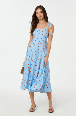 A woman is wearing a light blue, sleeveless, midi-length dress with thin shoulder straps tied into bows on her shoulders. The dress features an all-over pattern of large, evenly spaced, orange polka dots. She is also wearing nude-colored, open-toe heels and carrying a small, light-colored handbag.