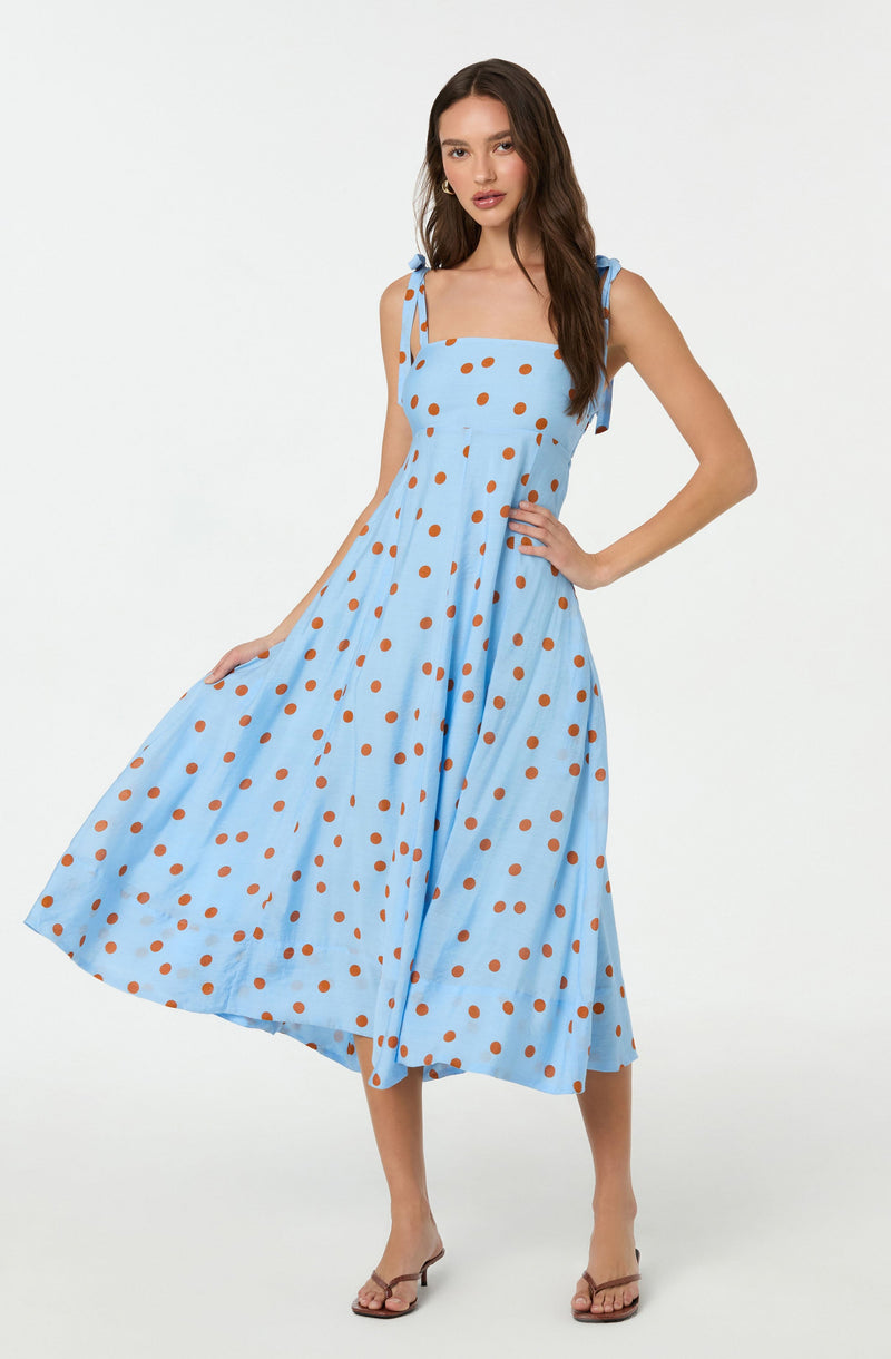 A woman is wearing a light blue, sleeveless, midi-length dress with thin shoulder straps tied into bows. The dress features a pattern of evenly spaced orange polka dots throughout. She is also wearing beige strappy sandals.