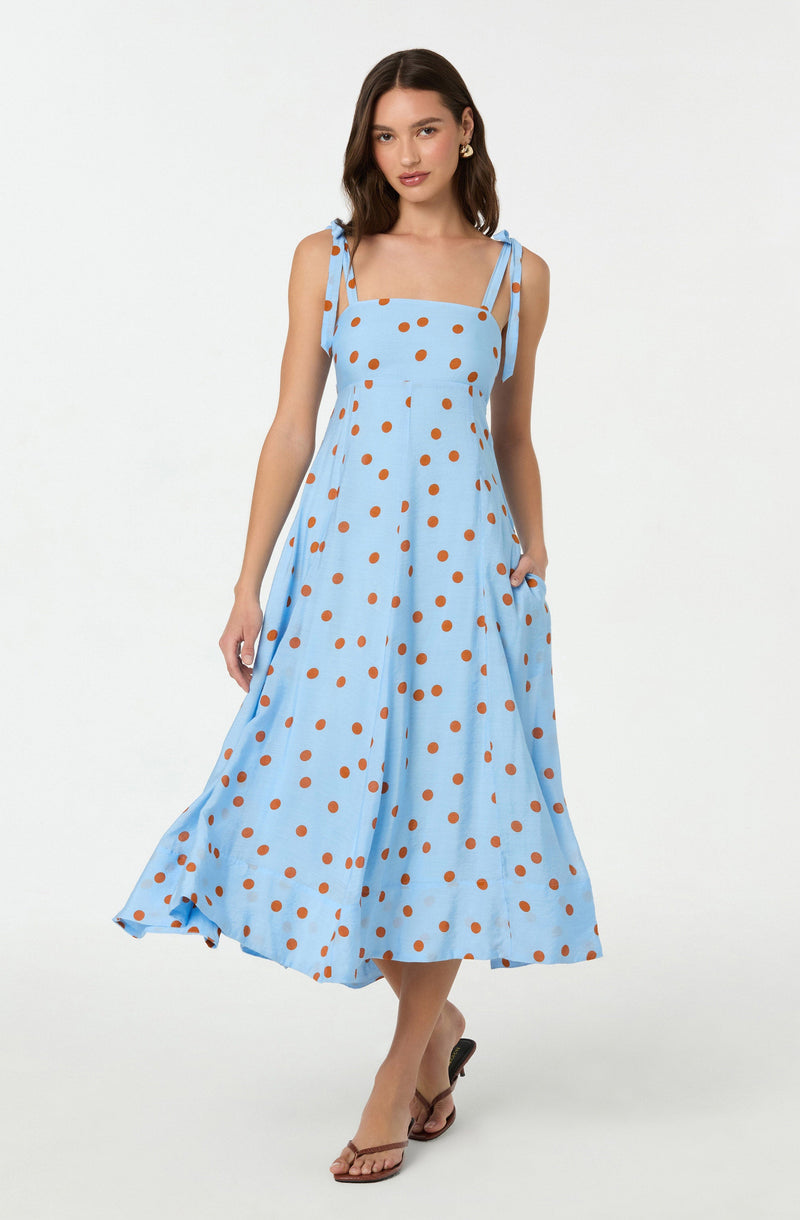 A woman is wearing a light blue sleeveless dress with thin, tied shoulder straps. The dress features a pattern of evenly spaced, large brown polka dots and has a fitted bodice with a gathered waist, flowing into a full, ankle-length skirt. She is also wearing tan open-toe sandals with a slight heel. The background is plain white.