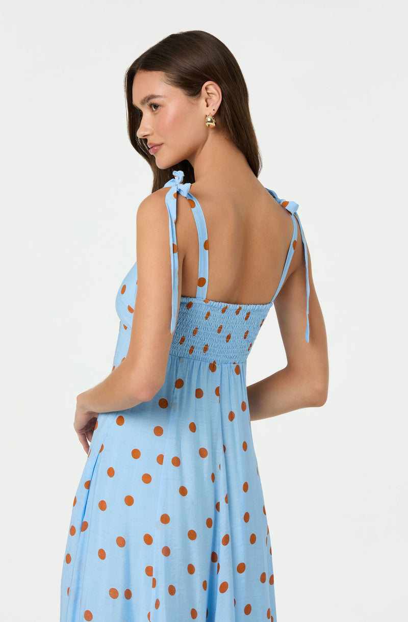 A woman is wearing a light blue sleeveless dress with brown polka dots. The dress has adjustable shoulder straps tied into bows and features a smocked waistline. The fabric appears lightweight and flowy.