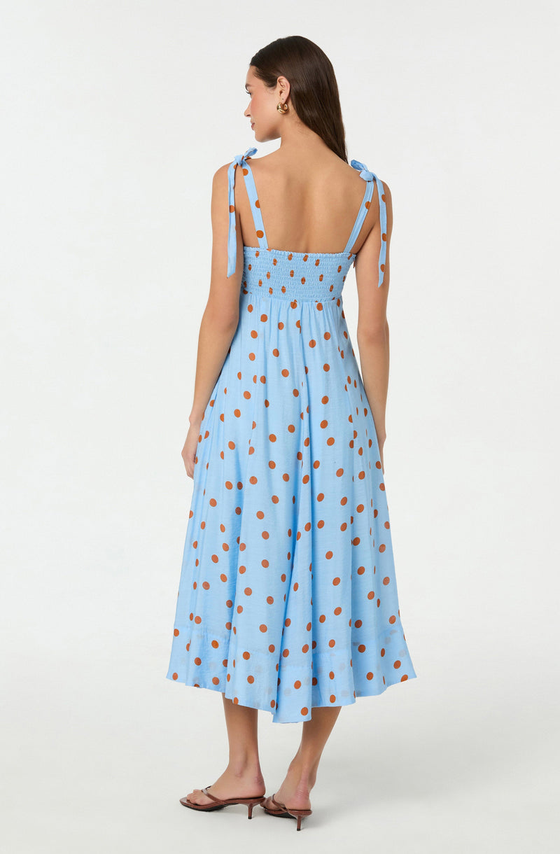 A woman is wearing a light blue midi dress with brown polka dots. The dress has shoulder straps that tie into bows on her shoulders, a gathered elasticized back, and a flowing skirt that reaches mid-calf length. She is paired with beige high-heeled sandals.