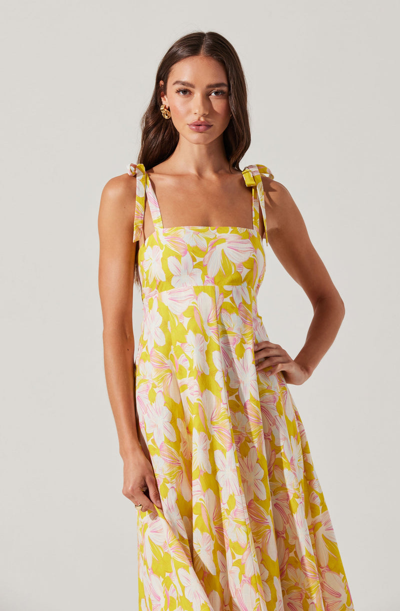A woman is wearing a sleeveless, ankle-length dress with a floral pattern of large white and pink flowers against a light yellow background. The dress features shoulder straps tied into bows.